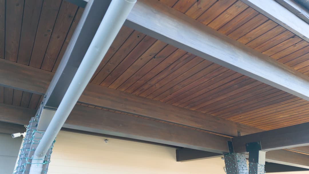 Wooden ceiling beams in a covered outdoor space with metal supports and grey drainage pipes.