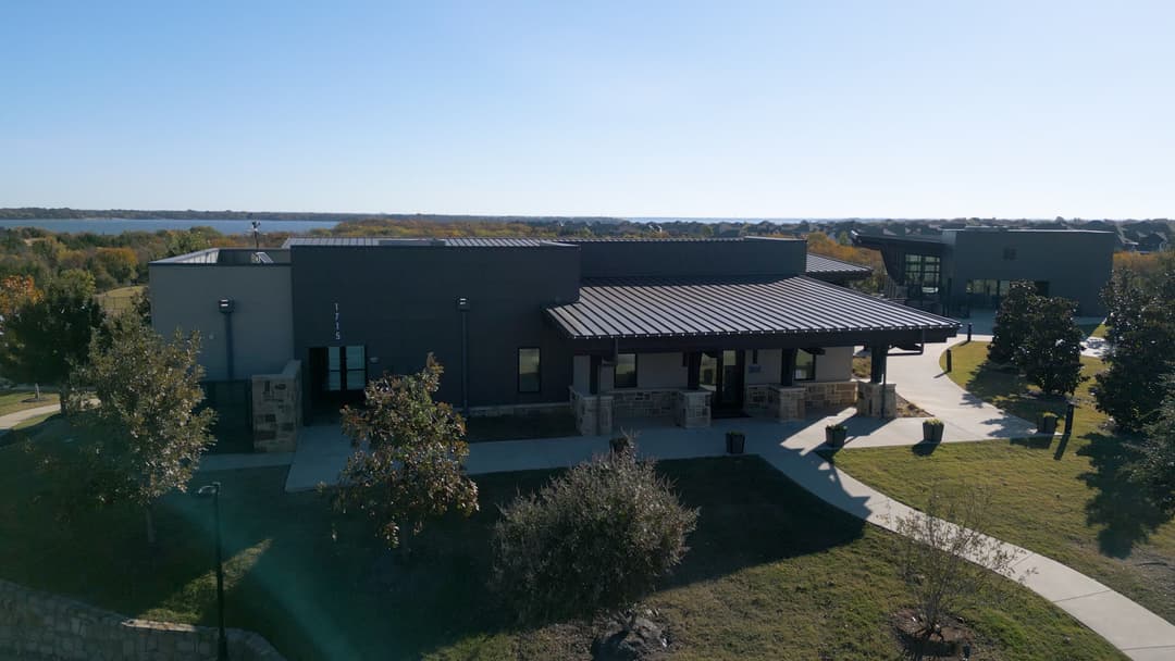 Modern community center with a metal roof surrounded by greenery and a lake view.