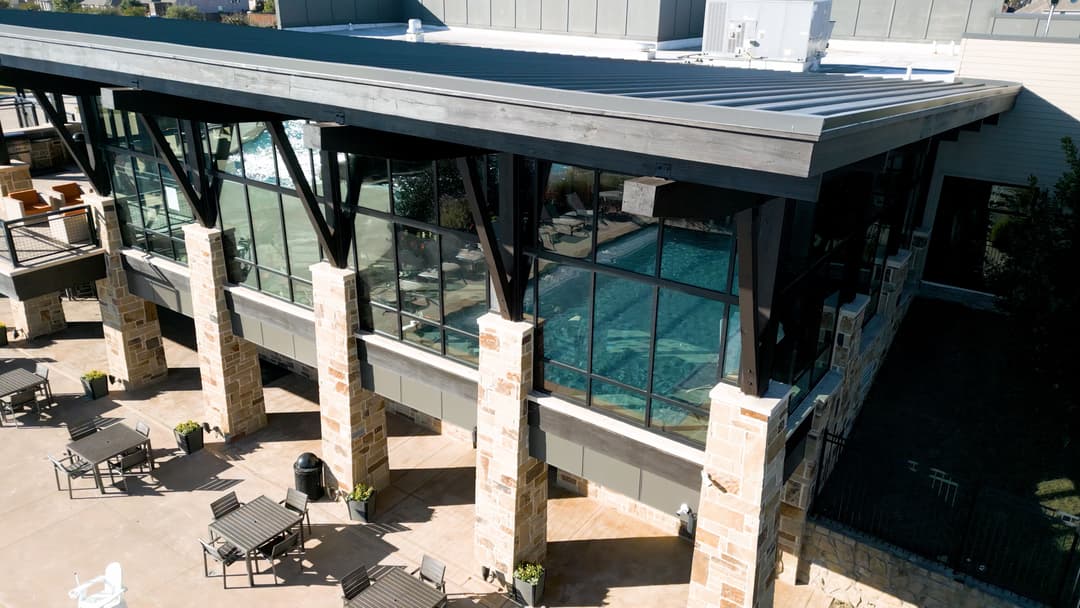 Modern building exterior with a large glass wall overlooking a pool area and patio seating.