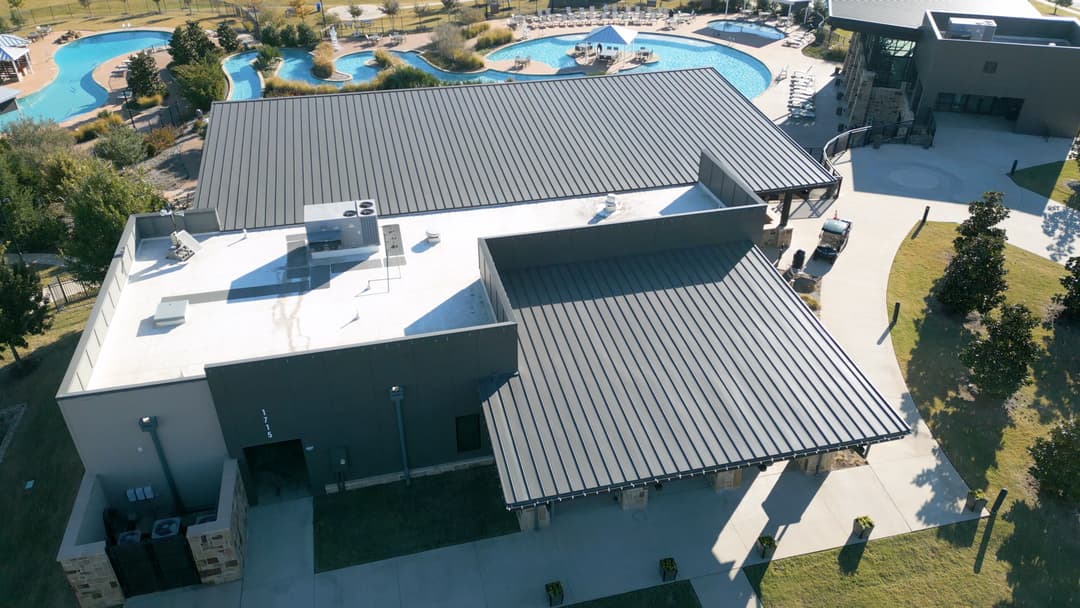 Aerial view of a modern building with a metal roof near a swimming pool and landscaped area.