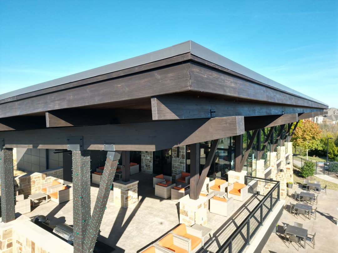 Modern restaurant patio with wooden beams, outdoor seating, and clear blue sky in the background.